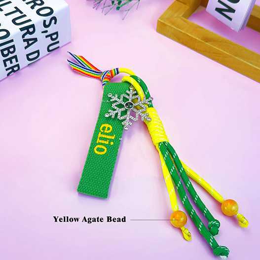 Green Christmas kids backpack charm with embroidered name “elio”, rhinestone snowflake, yellow agate beads, and rainbow ribbon on pink background.