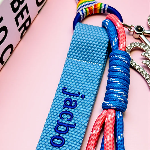 Close-up of blue cotton webbing name tag with embroidered letters and wrapped paracord on kids backpack charm.